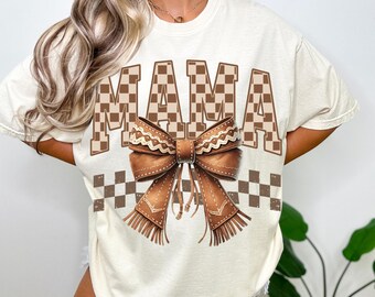 May include: A white t-shirt with a brown checkered pattern and the word "MAMA" in brown checkered letters. A brown bow with fringe is centered below the word.