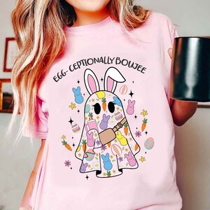 Puede incluir: Camiseta rosa con un fantasma con orejas de conejo y sosteniendo una bebida. El fantasma está rodeado de gráficos de temática de Pascua, incluyendo zanahorias, huevos y flores. El texto "Egg-ceptionally Boujee" está impreso en la camiseta.