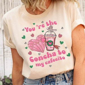 May include: A light beige t-shirt with a pink graphic design featuring a heart-shaped concha pastry and a cup of coffee with a straw. The text "You're the Concha to my cafecito" is written in pink letters.
