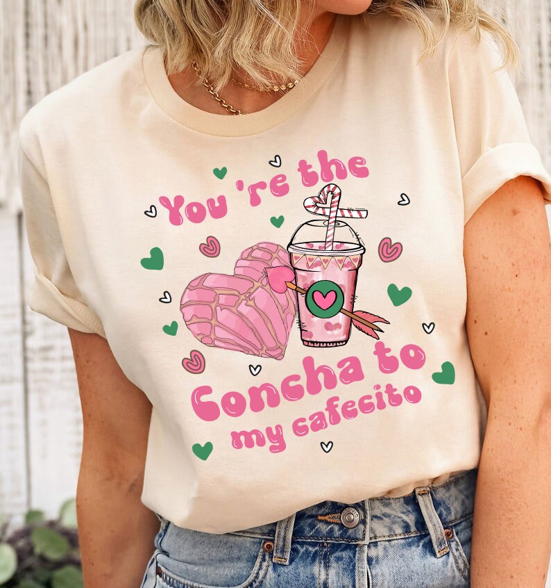 May include: A light beige t-shirt with a pink graphic design featuring a heart-shaped concha pastry and a cup of coffee with a straw. The text "You're the Concha to my cafecito" is written in pink letters.