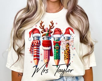 Puede incluir: Camiseta color crema con un diseño festivo de lápices. Cada lápiz está decorado con elementos navideños como gorros de Papá Noel, cuernos de reno y bufandas. El nombre "Mrs. Taylor" está escrito en cursiva debajo, lo que lo convierte en un regalo personalizado para profesores.