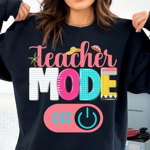 Puede incluir: Sudadera negra con el texto "Teacher Mode Off" en letras de colores. La palabra "Mode" está en una fuente grande y colorida con un gráfico de botón de encendido.