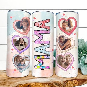May include: Three stainless steel tumblers with pink, white, and blue designs. Each tumbler features a heart-shaped frame with a photo of a mother and child. The middle tumbler has the word "MAMA" written in pink, white, and blue letters.