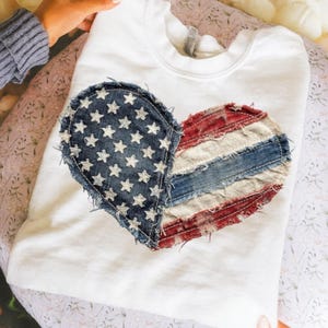May include: White sweatshirt featuring a heart-shaped design made from denim fabric. The left side of the heart displays a blue denim section with white stars, while the right side has red and blue stripes, resembling the American flag.