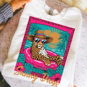 May include: White sweatshirt featuring a leopard wearing sunglasses and a straw hat, floating on a pink inflatable ring. The graphic includes a tropical scene with palm trees, a sunset, and the words "Sunny Days" in gold script.