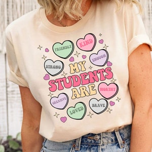 May include: A light beige t-shirt with a graphic design of candy hearts that say "My Students Are" and words like "Kind", "Strong", "Creative", "Loved", "Brave", "Worthy", "Enough", and "Friendly".