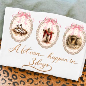 May include: White t-shirt featuring three oval designs with pink bows. The designs depict nails in a crown of thorns, a cross with a red cloth, and an empty tomb. The text "A lot can happen in 3 days" is written in gold script.