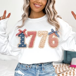 May include: White long-sleeve sweatshirt with the number "1776" appliqued on the front. The numbers are made of various patriotic fabrics, including blue with white stars, red gingham, floral, and red and white stripes. Each number is trimmed with lace and topped with a bow.