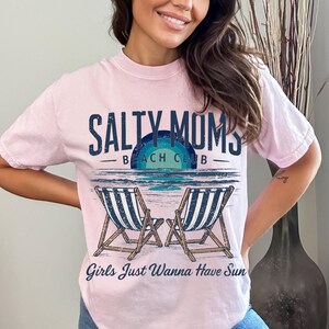 May include: Pink t-shirt with a graphic design that says "Salty Moms Beach Club" with two beach chairs and the text "Girls Just Wanna Have Sun".