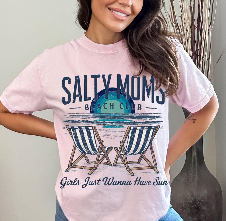 May include: Pink t-shirt with a graphic design that says "Salty Moms Beach Club" with two beach chairs and the text "Girls Just Wanna Have Sun".
