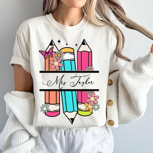 Könnte beinhalten: Weißes T-Shirt mit einem Design aus drei bunten Bleistiften mit Blumen und einem Schmetterling. Der Text "Mrs. Taylor" ist auf einem Banner zwischen den Bleistiften geschrieben.