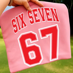 May include: A pink pillow with the words "SIX SEVEN" in white-outlined red letters and the number "67" in red with a white outline. The pillow is held up by a hand.