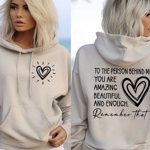 May include: A beige hoodie with a black heart design on the front and the text "TO THE PERSON BEHIND ME YOU ARE AMAZING BEAUTIFUL AND ENOUGH. Remember that" on the back.