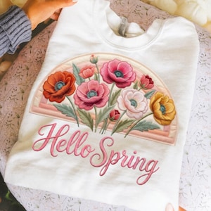 以下が含まれることがあります： 白いスウェットシャツ。花柄のデザイン、虹のアーチ、ピンクのスクリプトで「Hello Spring」の文字が描かれています。 花の配置には、オレンジ、ピンク、黄色の花と緑の葉が含まれています。 スウェットシャツは折りたたまれています。