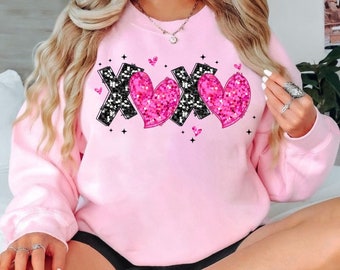 May include: A light pink sweatshirt with the letters "XOX" in a black glitter design and two pink glitter hearts. Small pink hearts and black stars are scattered around the design. The sweatshirt is a crew neck style.