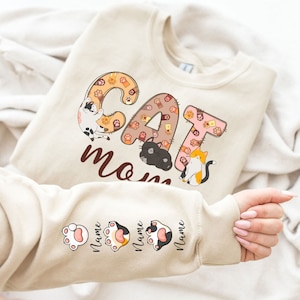 May include: Beige sweatshirt with the text "CAT MOM" in brown letters. The letters are made up of paw prints and cats. The sleeves have paw prints with the word "Name" written below each paw print.