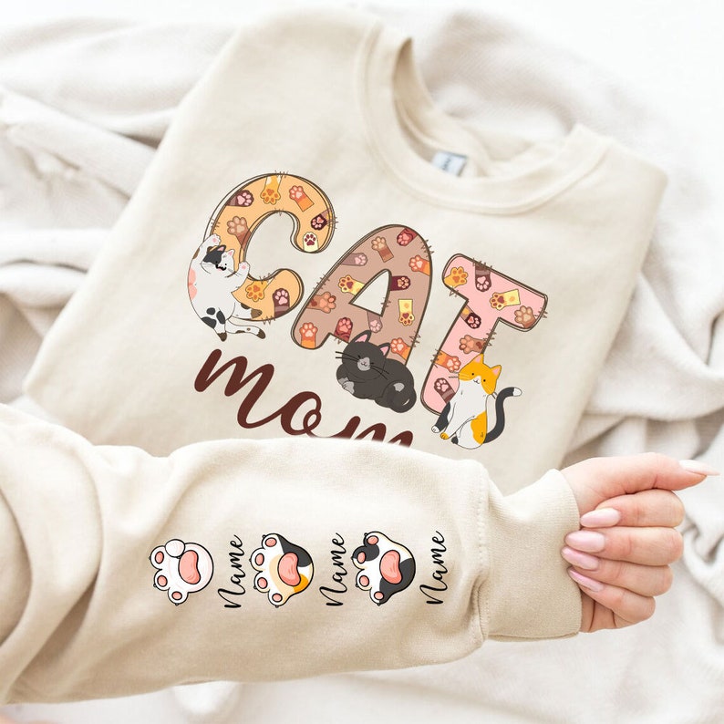 May include: Beige sweatshirt with the text "CAT MOM" in brown letters. The letters are made up of paw prints and cats. The sleeves have paw prints with the word "Name" written below each paw print.