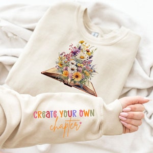 Puede incluir: Una sudadera beige con un ramo de flores que brota de un libro abierto. El texto "Crea tu propio capítulo" está impreso en la manga con letras de colores.