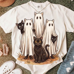 May include: A white short-sleeved t-shirt featuring a graphic print of three white ghost-costumed cats and two black cats. The central black cat sits in front of the ghosts, while the others peek out from behind the white fabric sheets.
