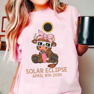 May include: Pink t-shirt with a cartoon brown cow wearing eclipse glasses and a pink bow. The cow is holding a pink drink. The text "SOLAR ECLIPSE APRIL 8TH 2024" is printed below the cow.