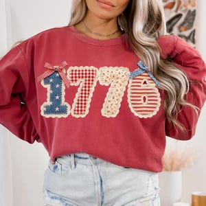 May include: A red sweatshirt with the number "1776" appliqued on the front. The numbers are made of various patriotic patterns, including stars, gingham, and stripes. Each number is outlined with a decorative stitch and topped with a bow.