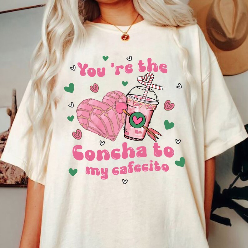 May include: A white t-shirt with a pink heart-shaped pastry and a pink cup of coffee with a straw. The text on the shirt reads "You're the Concha to my cafecito".
