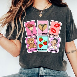 May include: A grey t-shirt with a pink and blue design featuring illustrations of a bouquet of flowers, tacos, lips, a teddy bear, strawberries, and Cupid. The text on the design reads "El ramo buch&oacute;n", "Los tacos", "Los besitos", "El oso de peluche", "Las fresas", and "El cupido".