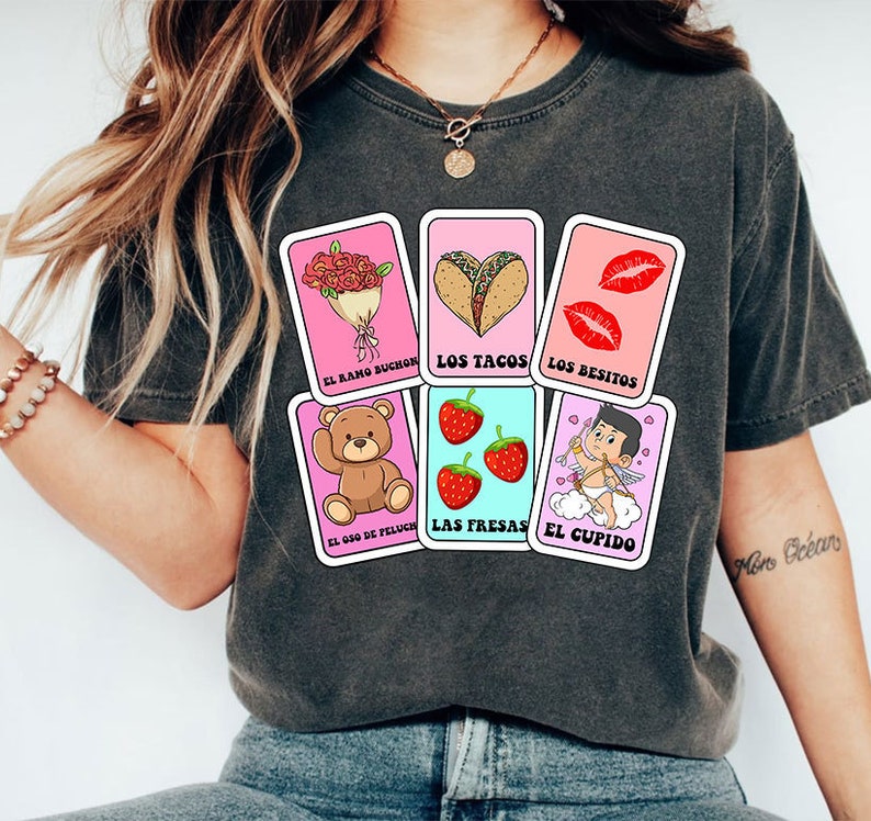 May include: A grey t-shirt with a pink and blue design featuring illustrations of a bouquet of flowers, tacos, lips, a teddy bear, strawberries, and Cupid. The text on the design reads "El ramo buch&oacute;n", "Los tacos", "Los besitos", "El oso de peluche", "Las fresas", and "El cupido".