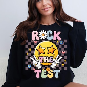Puede incluir: Sudadera negra con un diseño gráfico de una cara sonriente con estrellas por ojos y lápices por cejas. El texto "Rock the Test" está escrito en letras de colores.