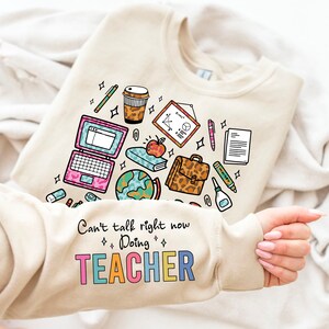 Puede incluir: Una sudadera beige con un diseño gráfico de útiles escolares, que incluye una computadora portátil, un globo terráqueo, una taza de café, un lápiz, una regla, un libro, una manzana, un maletín, un pegamento, una grapadora y un bloc de notas. El texto "Can't talk right now Doing TEACHER" está impreso debajo del gráfico.