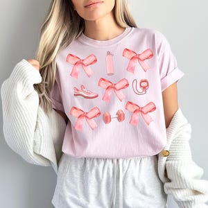 May include: Pink t-shirt featuring a graphic design of pink bows, a water bottle, a pair of running shoes, a dumbbell, and a heart rate monitor.