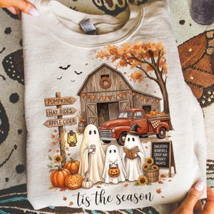 Puede incluir: Una sudadera beige de cuello redondo con un gráfico de un granero rústico, una camioneta roja, tres fantasmas, calabazas y hojas de otoño. El texto dice PUMPKINS, HAY RIDES, APPLE CIDER, SWEATERS, BONFIRES, CRISP AIR, SPOOKY NIGHTS y tis the season.