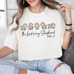 Pode incluir: Uma camiseta branca com um gráfico de uma linha de ovelhas e o texto "The Lord is my Shepherd Psalm 23:1"
