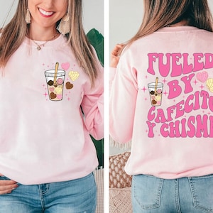 May include: Pink sweatshirt with a graphic of a coffee cup with pastries and the text "Fueled by Cafecito y Chisme" in pink letters.