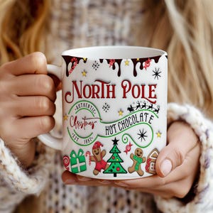 May include: White ceramic mug with a red, green and black design. The mug features the text "North Pole Authentic Christmas Hot Chocolate Est. 1997" with a gingerbread man, candy canes and a Christmas tree.