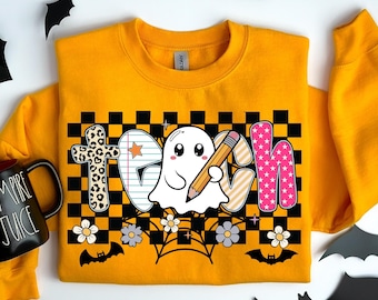 May include: A yellow sweatshirt featuring the word "Teach" in a graphic design. The design includes a ghost holding a pencil, a cross with a leopard print, and the letter "H" with pink stars. The design is set against a black and white checkered background.