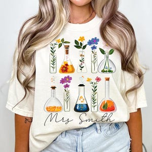 Pode incluir: Uma camiseta branca com um design floral com flores coloridas em béqueres. O texto "Mrs. Smith" é impresso abaixo dos béqueres.