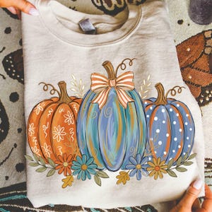 Puede incluir: Una sudadera de cuello redondo color beige con un gráfico de tres calabazas. La calabaza izquierda es naranja con patrones florales blancos, la central es azul con un lazo a rayas y la derecha es azul con lunares blancos, decoradas con flores.