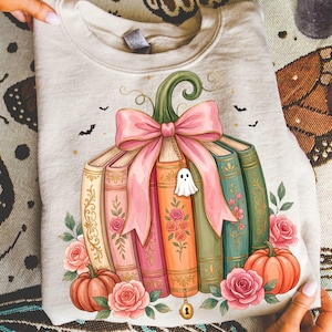 May include: A light beige crewneck sweatshirt featuring a graphic of a pumpkin shape formed by a stack of colorful vintage-style books. The books are tied with a large pink bow and a small ghost charm, surrounded by orange pumpkins and pink roses.