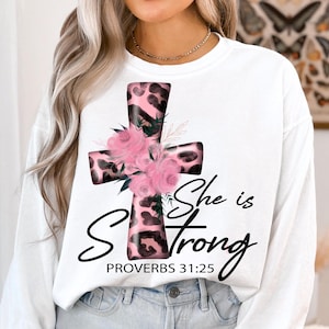 May include: White long-sleeved shirt with a pink and black leopard print cross with pink flowers and the text "She is Strong" and "Proverbs 31:25".