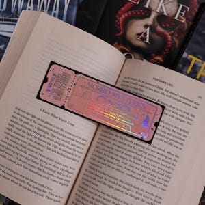 May include: A close-up of an open book with a pink and purple holographic bookmark. The bookmark has the words "Morbid Mysteries" and "I Know What You've Done" printed on it. The book is open to a page with text about a character named Jack.