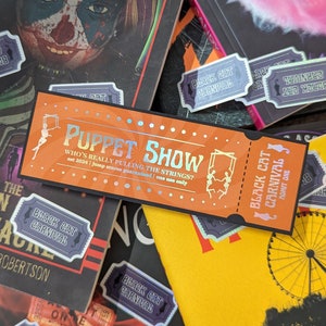May include: An orange ticket with the words "PUPPET SHOW" in holographic foil, with the text "WHO'S REALLY PULLING THE STRINGS?" and "est 2024 | jump scares guaranteed | one use only". Surrounding the ticket are books and "Black Cat Carnival" stickers.