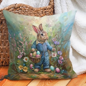Op de afbeelding: Een kussen met een schilderij van een konijn dat een overall draagt en een mand met paaseieren vasthoudt. Het konijn wordt omringd door bloemen en groen. De achtergrond is lichtblauw.