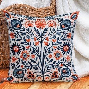 Op de afbeelding: Een decoratief kussen met een bloemenpatroon in tinten blauw, oranje en wit. Het kussen heeft een symmetrisch ontwerp met een centrale stengel en uitlopende bladeren.