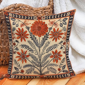 Op de afbeelding: Een crèmekleurig kussen met een bloemenpatroon. De bloemen zijn oranje en rood met zwarte stelen en bladeren. Het kussen heeft een zwart-witte rand.