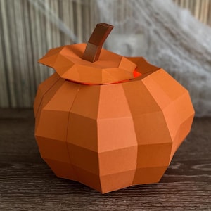 Può includere: Una zucca di carta arancione geometrica con un gambo marrone. La zucca ha un coperchio leggermente socchiuso, rivelando uno scorcio dell'interno. Questa zucca decorativa è perfetta per Halloween.