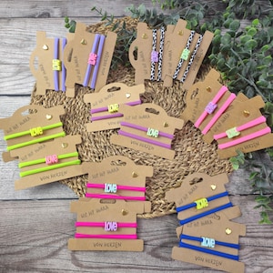Könnte beinhalten: Eine Sammlung bunter Haargummis, die auf Pappkarten präsentiert werden. Die Haargummis sind in verschiedenen Farben erhältlich, darunter Pink, Blau, Grün und Lila, mit dem Wort "Love" oder anderen Designs. Die Karten tragen den Text "Hip Hip Hurra" und "Von Herzen".