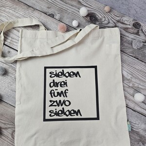 Könnte beinhalten: Eine beige Canvas-Tragetasche mit schwarzen Henkeln und einem schwarzen Quadrat auf der Vorderseite. Das Quadrat enthält die Wörter "sieben", "drei", "fünf", "zwei" und "sieben" in schwarzer, handschriftlicher Schrift.