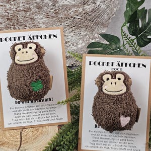 Könnte beinhalten: Zwei braune Plüschaffen auf weißen Präsentationskarten mit der Aufschrift POCKET ÄFFCHEN. Ein Affe hat einen grünen vierblättrigen Kleeblatt-Aufnäher, der andere, beschriftet mit Coco, einen rosa Herz-Aufnäher. Beide Karten enthalten deutschen Text.