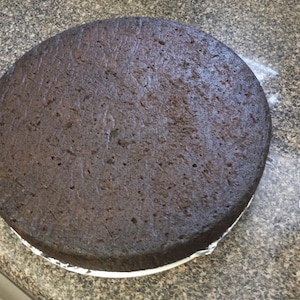 May include: A round, dark brown cake with a slightly textured surface. The cake is on a white paper liner and a gray countertop.